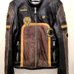 Könnte beinhalten: Eine schwarze und braune Lederjacke im Used-Look mit Reißverschluss und mehreren Aufnähern. Die Jacke hat braune und gelbe Akzente, mit Text wie "CAN SHAFT" und "SAKAWA".