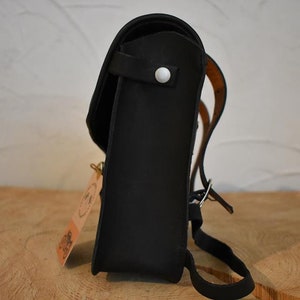May include: A black leather backpack with a white button on the front flap. The backpack has a brown leather strap and a buckle.
