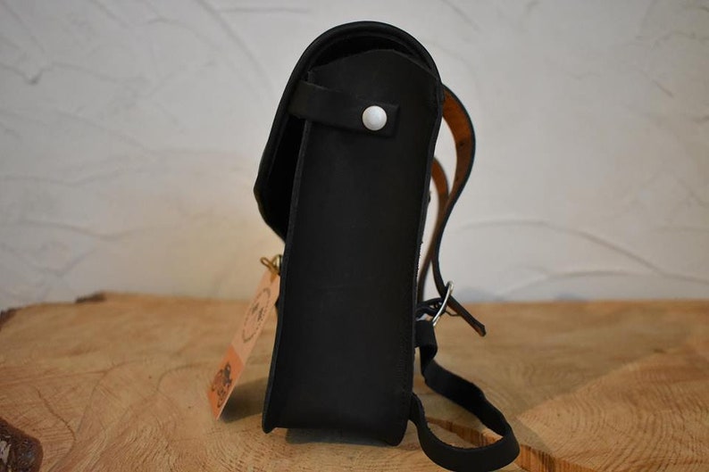 May include: A black leather backpack with a white button on the front flap. The backpack has a brown leather strap and a buckle.