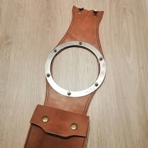 May include: A brown leather motorcycle sissy bar with a metal ring and a small pocket. The sissy bar is designed to be attached to a motorcycle seat.