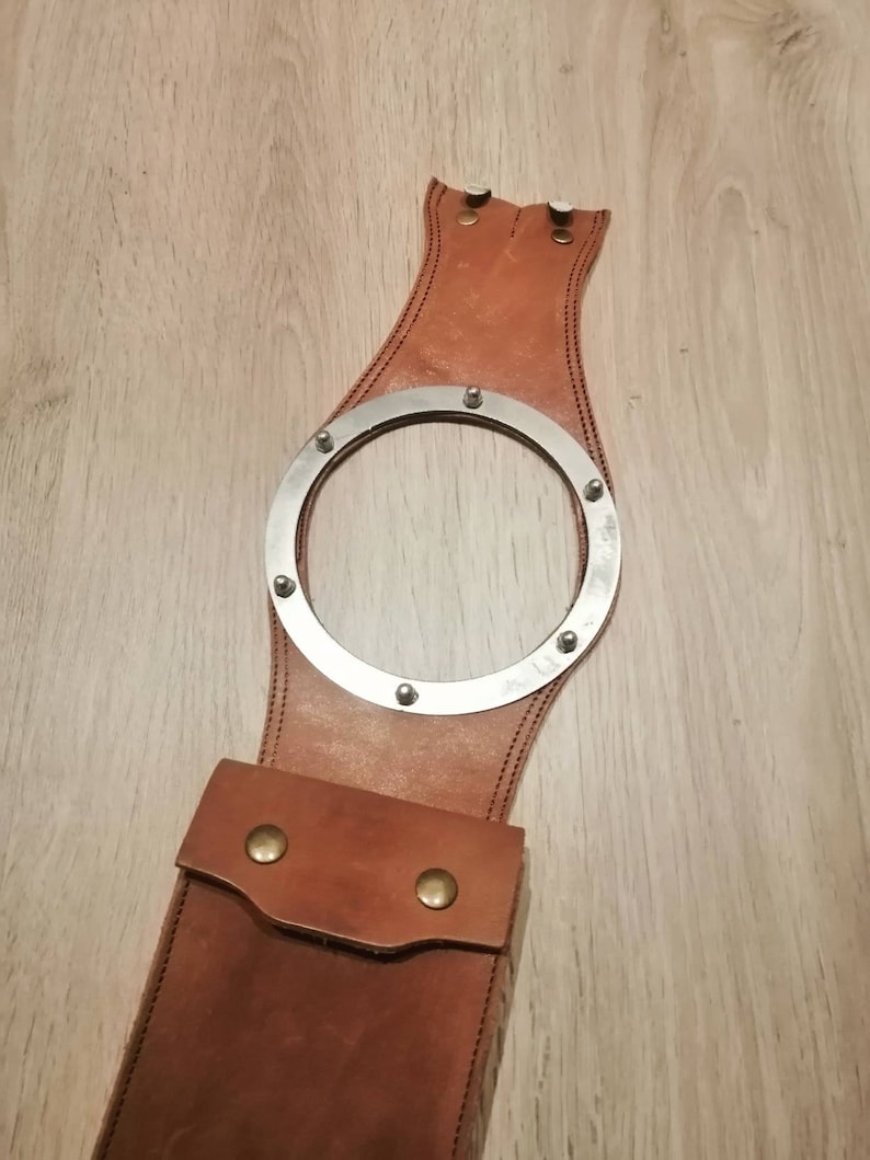 May include: A brown leather motorcycle sissy bar with a metal ring and a small pocket. The sissy bar is designed to be attached to a motorcycle seat.