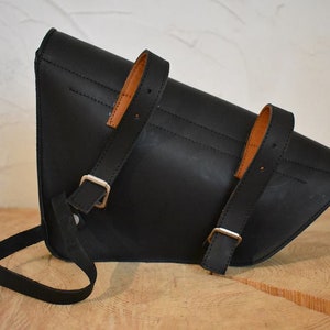May include: Black leather motorcycle saddlebag with brown leather straps and silver buckles. The bag has a triangular shape and a long strap for carrying.