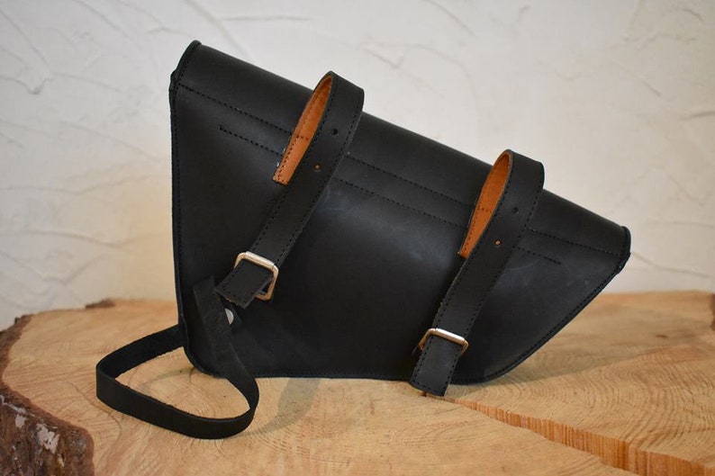 May include: Black leather motorcycle saddlebag with brown leather straps and silver buckles. The bag has a triangular shape and a long strap for carrying.