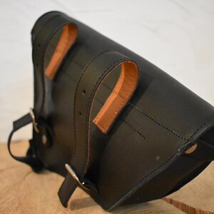 May include: Black leather motorcycle saddlebag with brown leather straps and buckles. The bag has a flap closure and a metal snap.