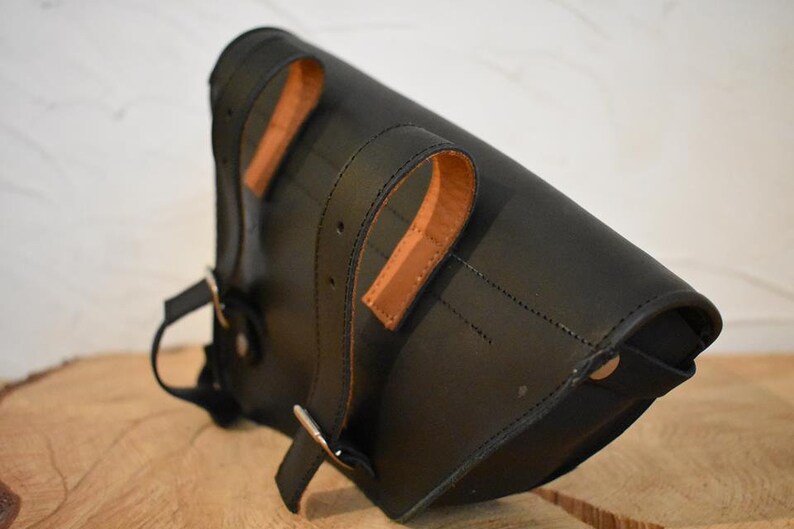 May include: Black leather motorcycle saddlebag with brown leather straps and buckles. The bag has a flap closure and a metal snap.