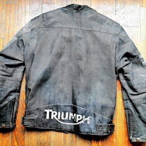 Könnte beinhalten: Graue Motorradjacke mit weißem Triumph-Logo auf dem unteren Rücken. Die Jacke hat einen Kragen, Reißverschlussärmel und ein Used-Look-Design. Die Jacke ist aus strapazierfähigem Material.