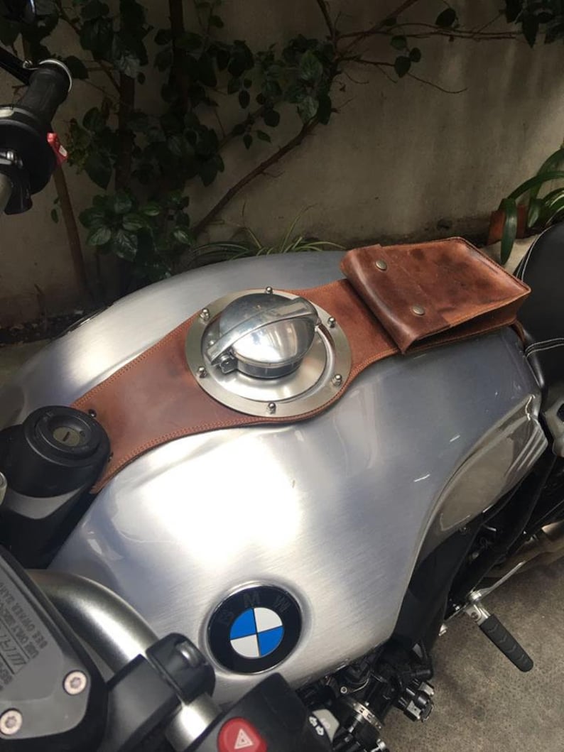 May include: A silver motorcycle gas tank with a brown leather strap and a small brown leather pouch. The tank has a round, silver gas cap with a BMW logo on the front.