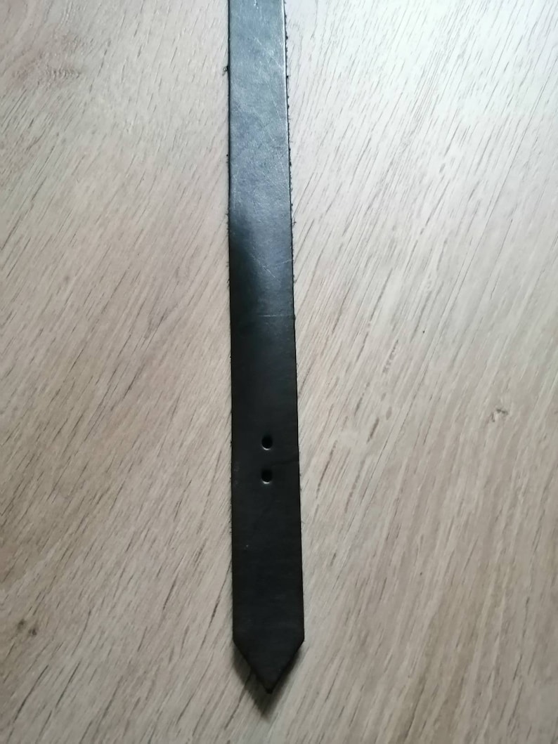 Puede incluir: Una correa de cuero negra con dos agujeros cerca del extremo, que se estrecha hasta un punto.