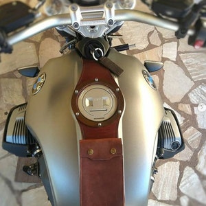 May include: A silver motorcycle gas tank with a brown leather strap and a round, silver gas cap. The strap has a pocket with two brass rivets.