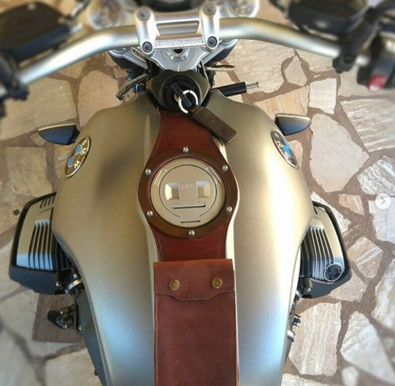 May include: A silver motorcycle gas tank with a brown leather strap and a round, silver gas cap. The strap has a pocket with two brass rivets.
