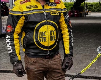May include: A black and yellow motorcycle jacket with the text "LUCKY STRIKE" on the front. The jacket has patches from "Michelin" and "Arrow". The person is wearing a matching black and yellow helmet and brown trousers.