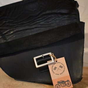 May include: Black leather motorcycle saddlebag with a silver buckle and a brown tag that says "Motard Classics and Riders Motorcycle".