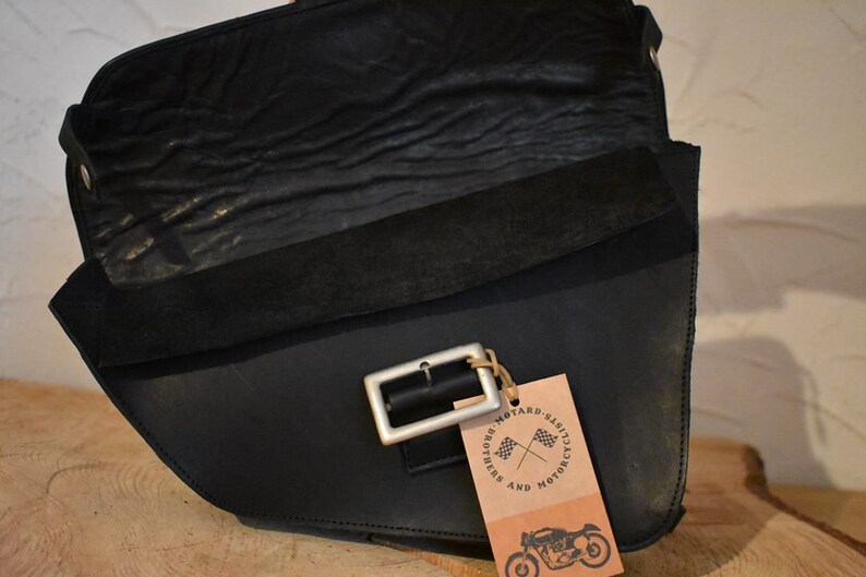May include: Black leather motorcycle saddlebag with a silver buckle and a brown tag that says "Motard Classics and Riders Motorcycle".