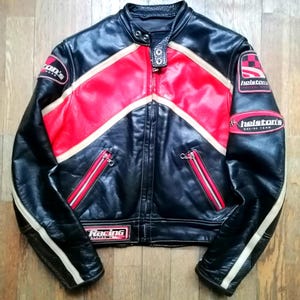 Può includere: Giacca da moto in pelle nera con dettagli rossi e color crema. Presenta un pannello centrale rosso e tasche con cerniera rosse. Include toppe con la scritta "helston's racing team" e "Racing".