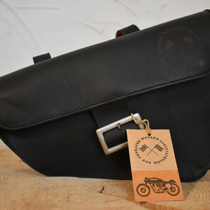 May include: Black leather motorcycle saddlebag with a silver buckle and a brown leather tag that says "And Riders' Motard's Classics" with a crossed checkered flag logo.