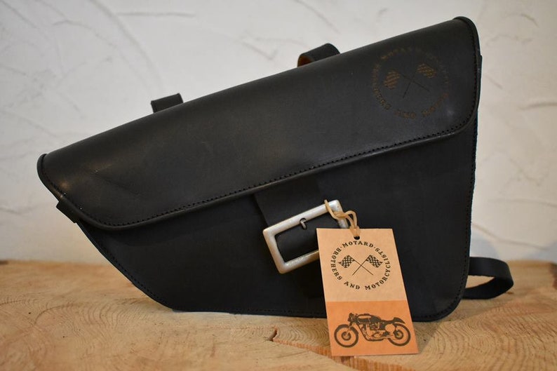 May include: Black leather motorcycle saddlebag with a silver buckle and a brown leather tag that says "And Riders' Motard's Classics" with a crossed checkered flag logo.
