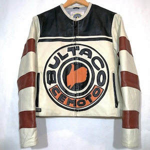 Könnte beinhalten: Weiße Leder-Motorradjacke mit einem schwarzen und orangefarbenen Bultaco Moto-Logo auf der Vorderseite. Die Jacke hat braun-weiße gestreifte Ärmel.