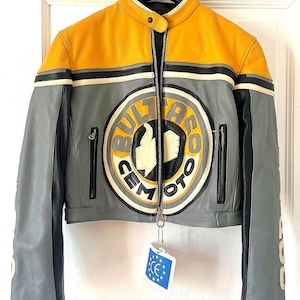 Könnte beinhalten: Grau, gelbe und schwarze Motorradjacke aus Leder mit einem "Bultaco Cemoto"-Logo. Die Jacke hat einen Reißverschluss, zwei Vordertaschen und ein Etikett mit CE-Symbol. Die Jacke hängt an einem Kleiderbügel.