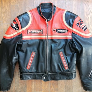 以下が含まれることがあります： 黒と赤の革製バイクジャケット。フロントジッパーと複数のポケット付き。ジャケットには、白とベージュのストライプと、「helston's racing team」と「Racing Collection」のテキストが入ったパッチが付いています。