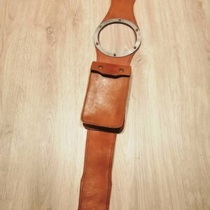 May include: A brown leather belt with a metal ring and a leather pouch. The belt is designed to be worn around the waist and has a metal ring that can be used to attach a key chain or other accessories. The pouch is attached to the belt and can be used to carry small items.