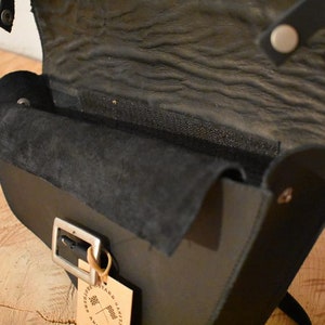May include: Black leather crossbody bag with a flap closure and a silver buckle. The bag has a suede lining and a small tag with the text "Artisan Crafted" on it.