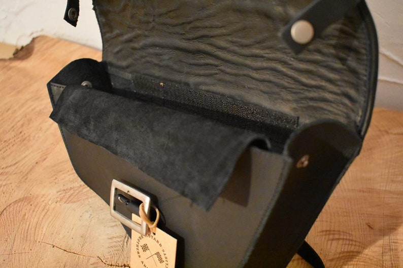 May include: Black leather crossbody bag with a flap closure and a silver buckle. The bag has a suede lining and a small tag with the text "Artisan Crafted" on it.