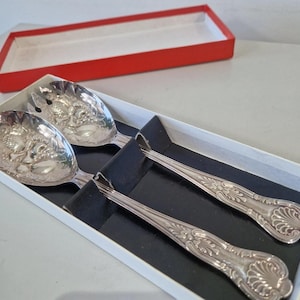 May include: A pair of silver-toned metal salad serving spoons with embossed fruit and leaf patterns on the bowls and decorative shell motifs on the handles. The utensils are presented in a white rectangular box with a black interior insert.