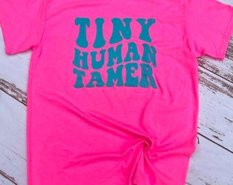 May include: A bright pink t-shirt with the text "TINY HUMAN TAMER" in turquoise blue lettering.