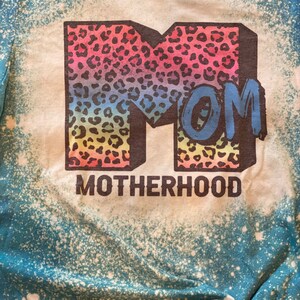 Op de afbeelding: Een blauw en wit tie-dye T-shirt met een regenboog luipaardprint "MOM" en de tekst "MOTHERHOOD" eronder.
