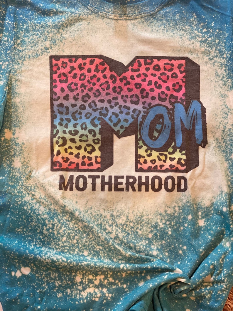 Op de afbeelding: Een blauw en wit tie-dye T-shirt met een regenboog luipaardprint "MOM" en de tekst "MOTHERHOOD" eronder.