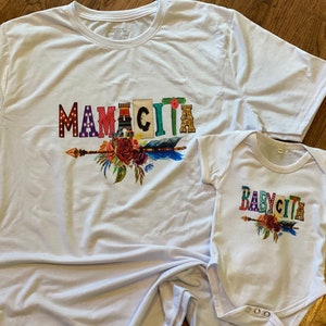 K&ouml;nnte beinhalten: Zwei wei&szlig;e T-Shirts mit einem bunten Grafikdesign. Das gr&ouml;&szlig;ere Shirt sagt "Mamacita" mit einem floralen Pfeil-Design. Das kleinere Shirt sagt "Babycita" mit einem &auml;hnlichen floralen Pfeil-Design.