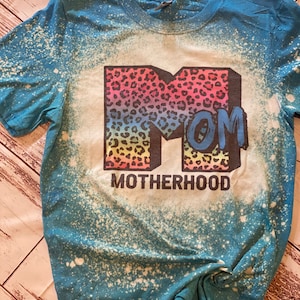 Op de afbeelding: Een turquoise blauw t-shirt met een ontwerp van bleekmiddelspatten. Het shirt heeft een grote, regenboogkleurige "M" met een luipaardprint. De "M" is omringd door het woord "MOM" en de tekst "MOTHERHOOD".