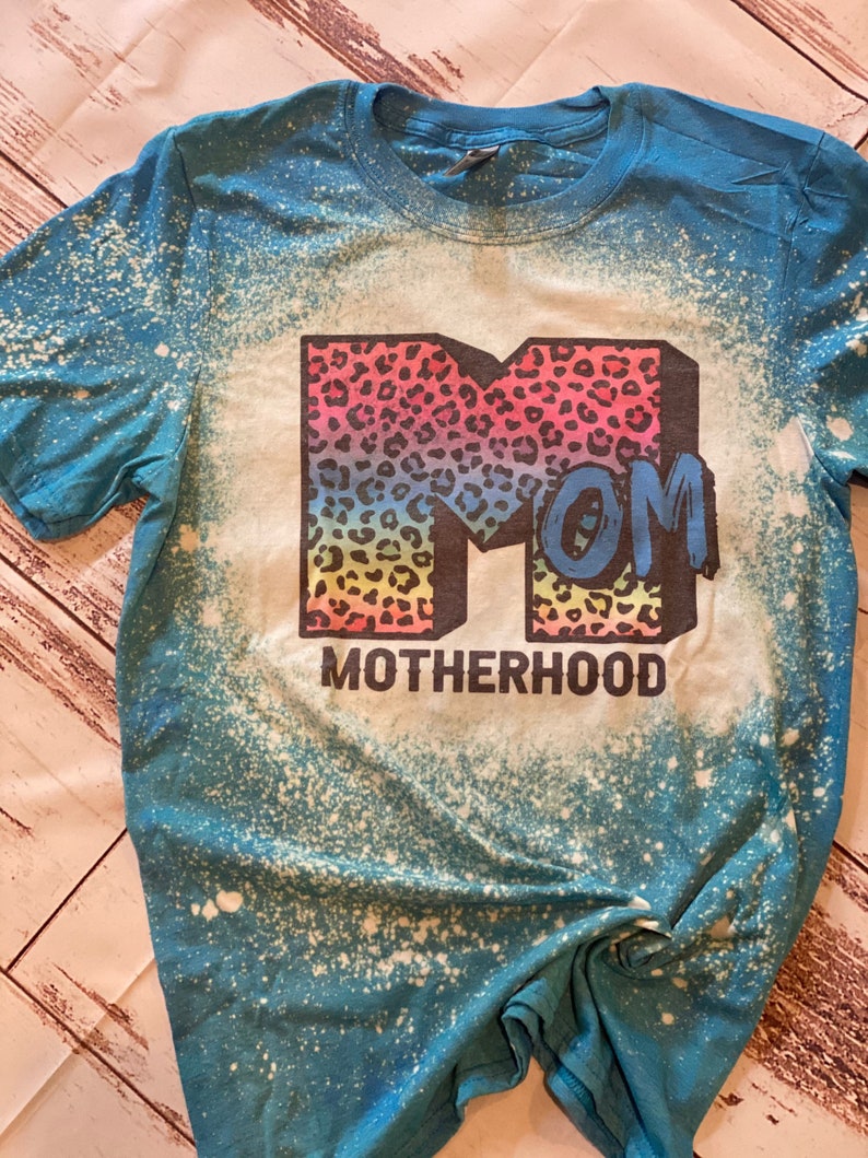 Op de afbeelding: Een turquoise blauw t-shirt met een ontwerp van bleekmiddelspatten. Het shirt heeft een grote, regenboogkleurige "M" met een luipaardprint. De "M" is omringd door het woord "MOM" en de tekst "MOTHERHOOD".
