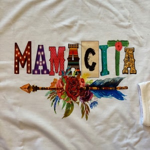 K&ouml;nnte beinhalten: Wei&szlig;es T-Shirt mit einem bunten Grafikdesign, das "Mamacita" mit einem floralen Pfeil-Design sagt.