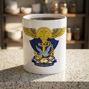May include: A white ceramic mug featuring a yellow and blue emblem with wings, an anchor, and a bird. The emblem includes the letters AC, the text SAR, and the motto SO OTHERS MAY LIVE printed in black at the bottom of the graphic design.