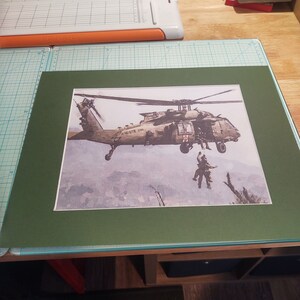 Pode incluir: Uma impress&atilde;o emoldurada de um helic&oacute;ptero militar em voo, com um trabalhador de resgate pendurado em uma corda. O helic&oacute;ptero &eacute; verde e cinza com as palavras "US Army" no lado. O fundo &eacute; uma aquarela de azul e cinza.
