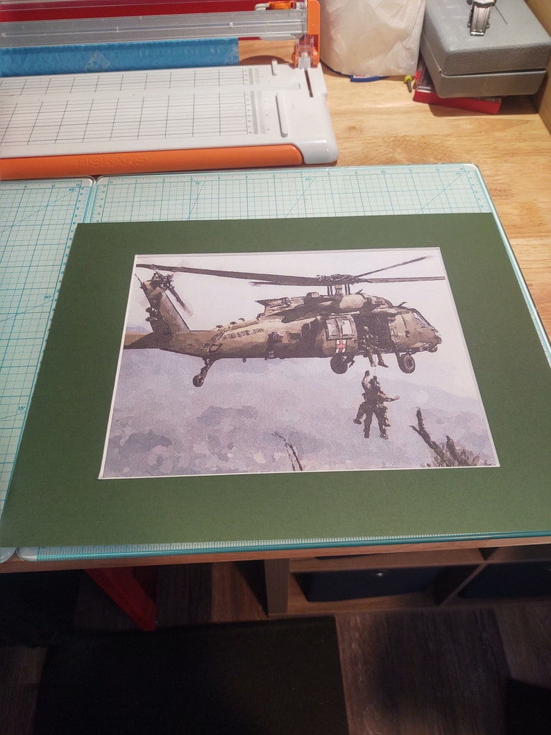 Pode incluir: Uma impress&atilde;o emoldurada de um helic&oacute;ptero militar em voo, com um trabalhador de resgate pendurado em uma corda. O helic&oacute;ptero &eacute; verde e cinza com as palavras "US Army" no lado. O fundo &eacute; uma aquarela de azul e cinza.