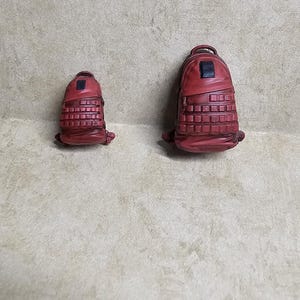 May include: Two miniature red backpacks of different sizes, each featuring a grid-like textured front panel, a small black rectangular accent near the top, a rounded main compartment, and a top handle, displayed side by side on a neutral surface.