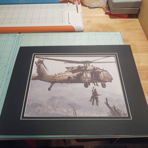Pode incluir: Uma impress&atilde;o emoldurada de um helic&oacute;ptero militar em voo, com dois soldados descendo de rappel da ba&iacute;a de carga aberta. O helic&oacute;ptero est&aacute; pintado em cores de camuflagem e tem uma cruz vermelha no lado. A impress&atilde;o &eacute; colocada sobre um fundo de c&eacute;u nublado e montanhas distantes.