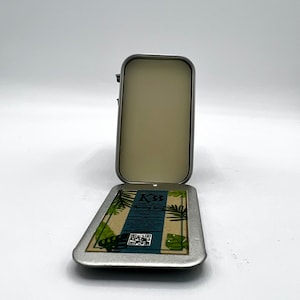 May include: An open, rectangular, silver-colored tin with a hinged lid. The inside of the tin is a light beige color. The bottom of the tin has a label with text and a QR code, with green and brown tropical leaf designs.