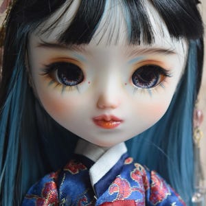 May include: A doll with black and blue hair, wearing a blue and red patterned traditional outfit. The doll has large eyes and a hair accessory. The text "IG: KITASIN_DOLLS" is visible on the image.