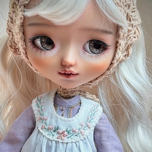 May include: A detailed close-up of a doll with a serene expression. The doll has large, expressive eyes, rosy cheeks, and a delicate mouth. It wears a light purple dress with a white bib adorned with floral embroidery and a crocheted bonnet.