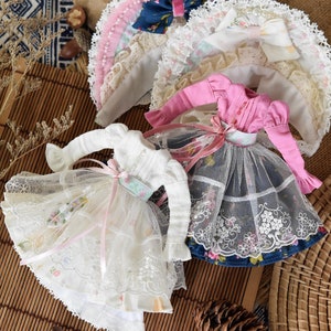 May include: Two doll dresses with lace trim and floral patterns. One dress is white with a pink ribbon and the other is pink with a blue and white floral print.