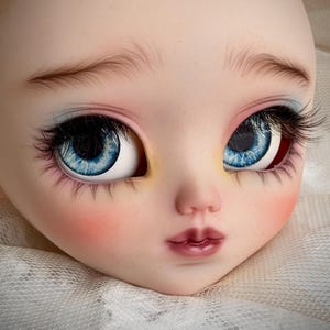May include: A doll head with a pale complexion, detailed with blue eyes, long black eyelashes, and delicate brown eyebrows. The cheeks are blushed pink, and the lips are a deep rose color. The head rests on a white, textured surface.
