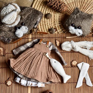 May include: A collection of handmade doll accessories, including a brown dress with suspenders, a plaid shirt, a white fish-shaped charm, and a plush animal head with a hat. The items are arranged on a bamboo mat.