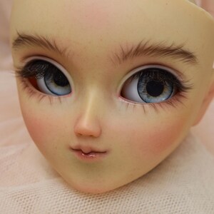 May include: A close-up of a doll's face with detailed features. The doll has light skin, blue eyes with long eyelashes, and delicately painted eyebrows. The lips are a soft pink, and the cheeks have a subtle blush. The text "IG: KITASIN_DOLLS_HOUSE" is at the bottom.