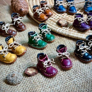 May include: A collection of miniature leather boots in various colors including brown, yellow, green, purple, and blue. Each tiny boot features white laces, visible stitching along the sole, and a rounded toe design, arranged on a textured surface.
