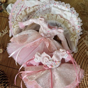 May include: Two pink doll dresses with lace and tulle. The top dress has a white bow and the bottom dress has a pink bow.