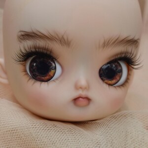 May include: A close-up of a doll's face with large, expressive eyes. The eyes are a striking mix of brown and amber, framed by long, dark eyelashes and delicate eyebrows. The doll has a pale complexion and a soft, neutral-toned background. The text "IG: KITASIN_DOLLS_HOUSE" is visible.
