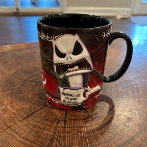 May include: Black ceramic mug featuring Jack Skellington from The Nightmare Before Christmas. The mug has a black handle and interior, with a design of Jack in a red and black background. Text on the mug reads "Mornings are my worst nightmare."